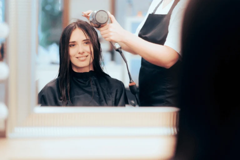Stylist begins blow-drying client's wet hair as she smiles in salon mirror reflection during appointment