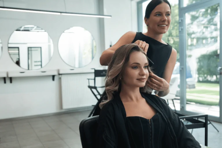Photo shows stylist arranging client’s curled hair in salon chair, blowout with extensions service