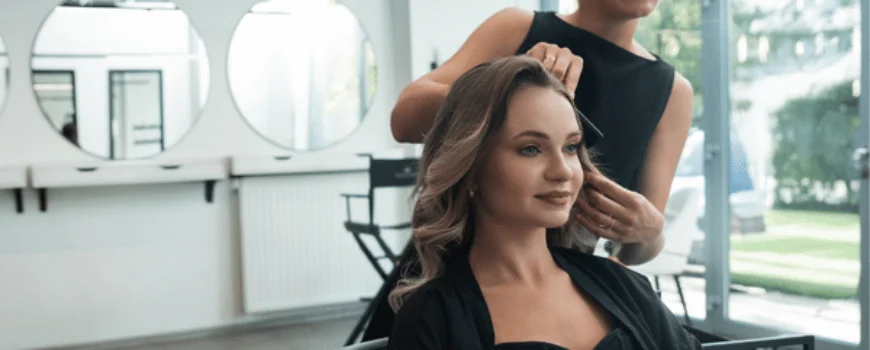 Photo shows stylist arranging client’s curled hair in salon chair, blowout with extensions service